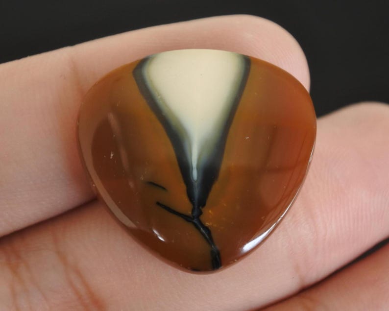 May include: A polished, heart-shaped gemstone with a glossy finish. The stone features a central ivory-colored wedge, bordered by a dark gray band, and encased in a rich, reddish-brown hue. A thin black vein-like pattern extends from the base.