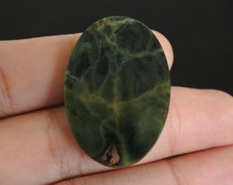 May include: An oval gemstone, predominantly dark green with lighter green and yellow veining. The polished stone is held in a hand, set against a dark backdrop. Suitable for jewellery, the stone's measurements would be in centimetres.