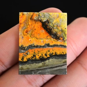 May include: A rectangular gemstone with a vibrant pattern of orange, yellow, black, and gray. The stone's surface displays a layered, textured appearance, with bands of color creating a unique visual effect. The gemstone is held in a hand.