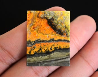 May include: A rectangular gemstone with a vibrant pattern of orange, yellow, black, and grey. The stone's surface displays a layered, textured appearance, with bands of colour creating a unique visual effect. The gemstone is held in a hand.