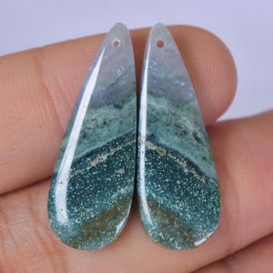 May include: Two teardrop-shaped gemstones with a hole at the top for jewelry making. The stones feature a mix of green, blue, and gray hues with a speckled pattern. The bottom portion of the stones has a darker, more saturated green color.