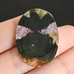 May include: Oval-shaped gemstone with a polished surface. The stone features a dark base color with patches of pink, green, and orange hues. The gemstone is held in a hand against a black background.