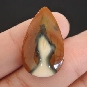May include: A polished, teardrop-shaped gemstone with a mix of colors. The stone features a base of creamy white, overlaid with a pattern of black and brown, and a glossy finish. The gemstone is held in a hand.