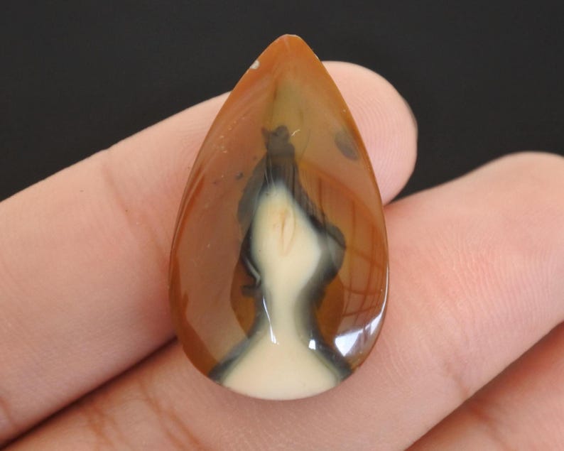 May include: A polished, teardrop-shaped gemstone with a mix of colours. The stone features a base of creamy white, overlaid with a pattern of black and brown, and a glossy finish. The gemstone is held in a hand.
