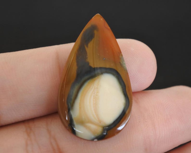 May include: A polished, teardrop-shaped gemstone. The stone displays a mix of brown, black, and cream hues, forming a distinctive natural pattern. The gemstone is held in a hand, highlighting its size and form. The stone's surface is smooth and reflective.