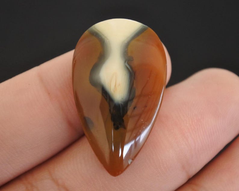 May include: A polished, teardrop-shaped gemstone with a glossy finish. The stone features a blend of cream, brown, and black, creating a unique pattern. The gemstone is held in a hand, showcasing its size and shape.