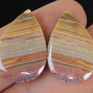 May include: Two teardrop-shaped gemstones with a polished surface. The stones display layers of tan, orange, and purple hues. The gemstones are held in a hand, with a dark background.