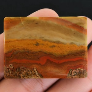 Op de afbeelding: Een rechthoekige steen met een gepolijst oppervlak. De steen heeft kleurlagen, waaronder tinten beige, oranje en rood. De onderste rand heeft een ruwe, getextureerde uitstraling, terwijl de bovenkant glad is.