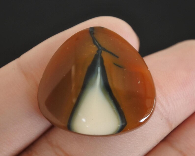 May include: A polished, triangular gemstone with a striking pattern. The stone features a blend of warm colors, including shades of brown and orange, with a central white and black design. The gemstone is held in a hand.