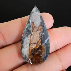 May include: A teardrop-shaped gemstone with a polished surface. The stone features a blend of colours, including grey, brown, white, and hints of blue. The pattern resembles a landscape with layered textures and colour variations.