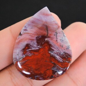 Puede incluir: Una gema pulida en forma de lágrima. La piedra presenta un llamativo patrón de tonos rojo intenso, blanco y morado, con vetas intrincadas y un acabado brillante. La gema se sostiene en una mano, mostrando su belleza natural.