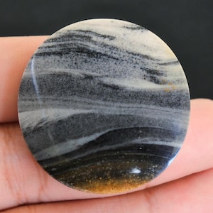 Op de afbeelding: Een ronde, gepolijste steen met een wervelend patroon van zwart, grijs, wit en bruin. Het oppervlak van de steen is glad en reflecterend, en toont de natuurlijke variaties in kleur en textuur. De steen wordt in een hand gehouden.