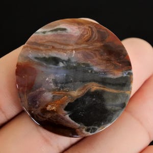 Peut inclure: Une pierre précieuse polie et ronde avec un motif tourbillonnant de teintes marron, noir, blanc et brun rougeâtre. La pierre a une finition brillante et un aspect naturel et organique. La pierre est tenue dans une main.
