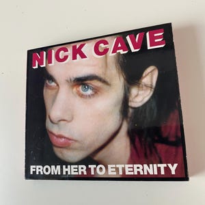 Peut inclure: Une pochette d'album en noir et blanc avec une photo rapprochée de Nick Cave. Le titre de l'album "From Her To Eternity" est imprimé en lettres blanches sur fond noir.