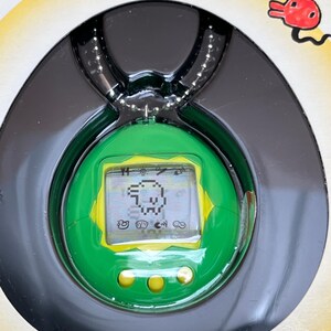 May include: A green and yellow Tamagotchi keychain with a digital screen displaying a small pixelated creature. The keychain is in its original packaging and has a silver chain.