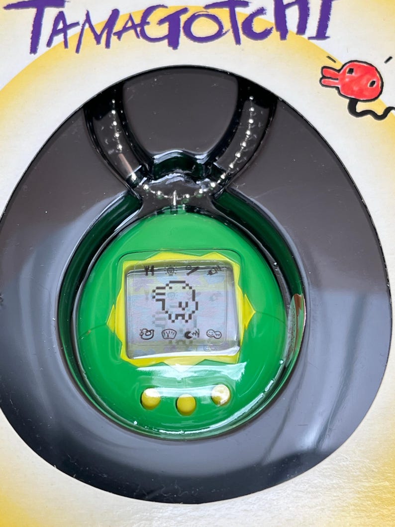 May include: A green and yellow Tamagotchi keychain with a digital screen displaying a small pixelated creature. The keychain is in its original packaging and has a silver chain.
