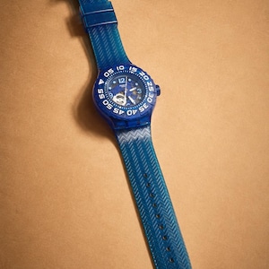 Puede incluir: Un reloj de pulsera azul con una correa transparente y una esfera azul. El reloj tiene una esfera redonda con números y agujas blancos. La correa tiene un patrón texturizado y se abrocha con una hebilla.