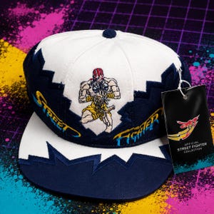 May include: A white and navy blue snapback cap featuring an embroidered Street Fighter character on the front panel. The word Street is embroidered in yellow and blue on the left, Fighter on the right. Includes an Official Street Fighter Collection tag.
