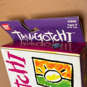 May include: A purple and white box with the text "Tamagotchi" on it. The box is for a Tamagotchi toy and has a picture of a sun on it. The box is labeled with the number "#1800" and says "Ages 8 and Up."