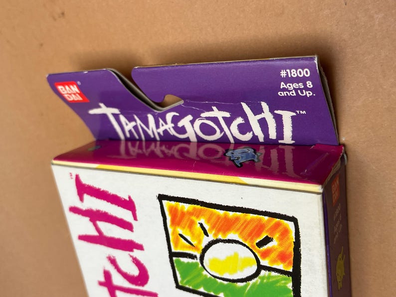 May include: A purple and white box with the text "Tamagotchi" on it. The box is for a Tamagotchi toy and has a picture of a sun on it. The box is labeled with the number "#1800" and says "Ages 8 and Up."