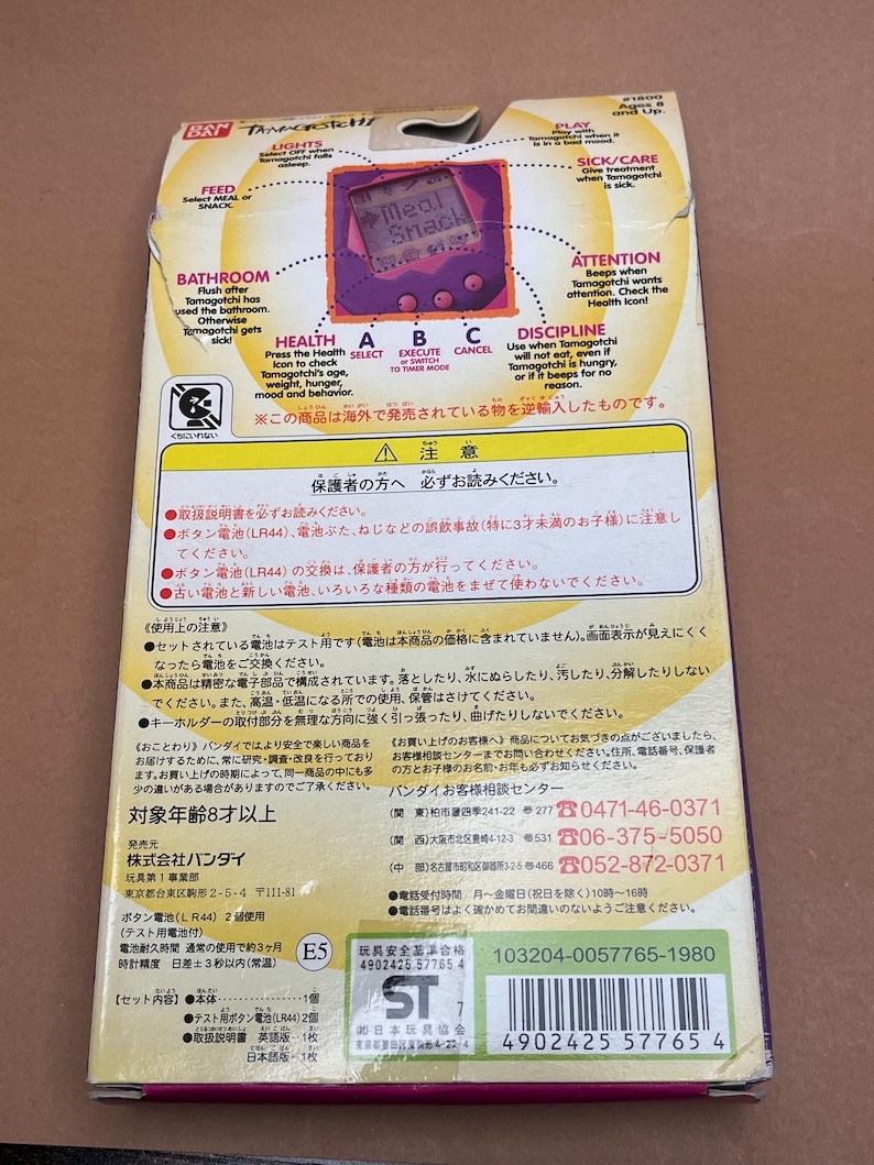 May include: A purple and pink Tamagotchi electronic pet toy with a screen that reads "Meal Snack". The box is pink and white with Japanese text and instructions. The box includes a warning to parents to read the instructions.