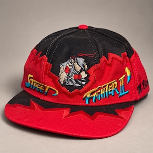 May include: A black and red baseball cap featuring an embroidered graphic of the character Ryu from Street Fighter II. The cap displays the Street Fighter II logo in yellow and blue, with a jagged red and black pattern and the text M. Bison.