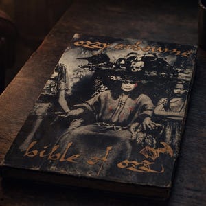 May include: A vintage book titled "Bible of Ozz" by Ozzy Osbourne, featuring a dark, gothic illustration on the cover. The cover art depicts a seated figure surrounded by other figures and skulls. The book rests on a dark wooden surface.