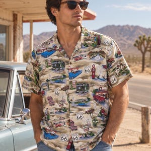 May include: A short-sleeved button-down shirt featuring a tan background with a colorful all-over print of Route 66 imagery, including vintage cars, motels, gas pumps, and road signs. The shirt has a classic collar and a front button closure.
