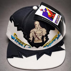May include: A black and white baseball cap featuring an embroidered muscular figure on the front panel. The cap displays the text Street Fighter in blue and yellow embroidery. A black tag with a Capcom logo and character illustration is attached.