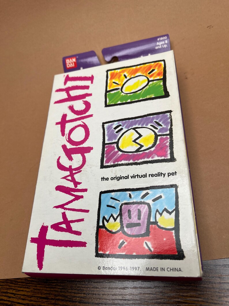 May include: A pink, white, and blue box with the words "Tamagotchi" in large pink letters. The box features three colorful illustrations of the Tamagotchi character in different stages of development. The text "the original virtual reality pet" is printed below the illustrations. The box also includes the text "Bandai 1996-1997, MADE IN CHINA."