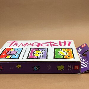May include: A white box with the words "Tamagotchi" in pink letters. The box has three colorful images of the Tamagotchi characters. The box is labeled "The original virtual pet" and "Ages 8 and Up". The box is also labeled "#1500" and "Bandai".