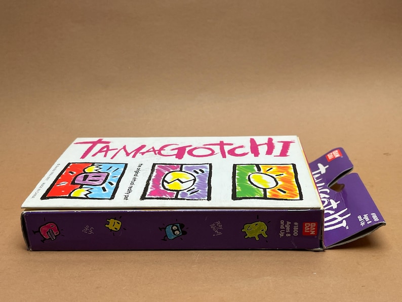 May include: A white box with the words "Tamagotchi" in pink letters. The box has three colorful images of the Tamagotchi characters. The box is labeled "The original virtual pet" and "Ages 8 and Up". The box is also labeled "#1500" and "Bandai".