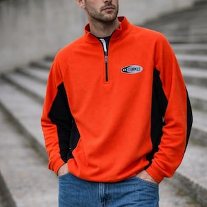 Op de afbeelding: Een oranje fleece trui met zwarte zijpanelen en een zwarte rits. De trui heeft de tekst "HOOKWILD" op de borst. De persoon draagt een blauwe spijkerbroek.