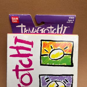May include: A purple and white box with the text "Tamagotchi" and "#1800 Ages 8 and Up." The box features a cartoon image of a yellow egg with a sun and a cracked egg.