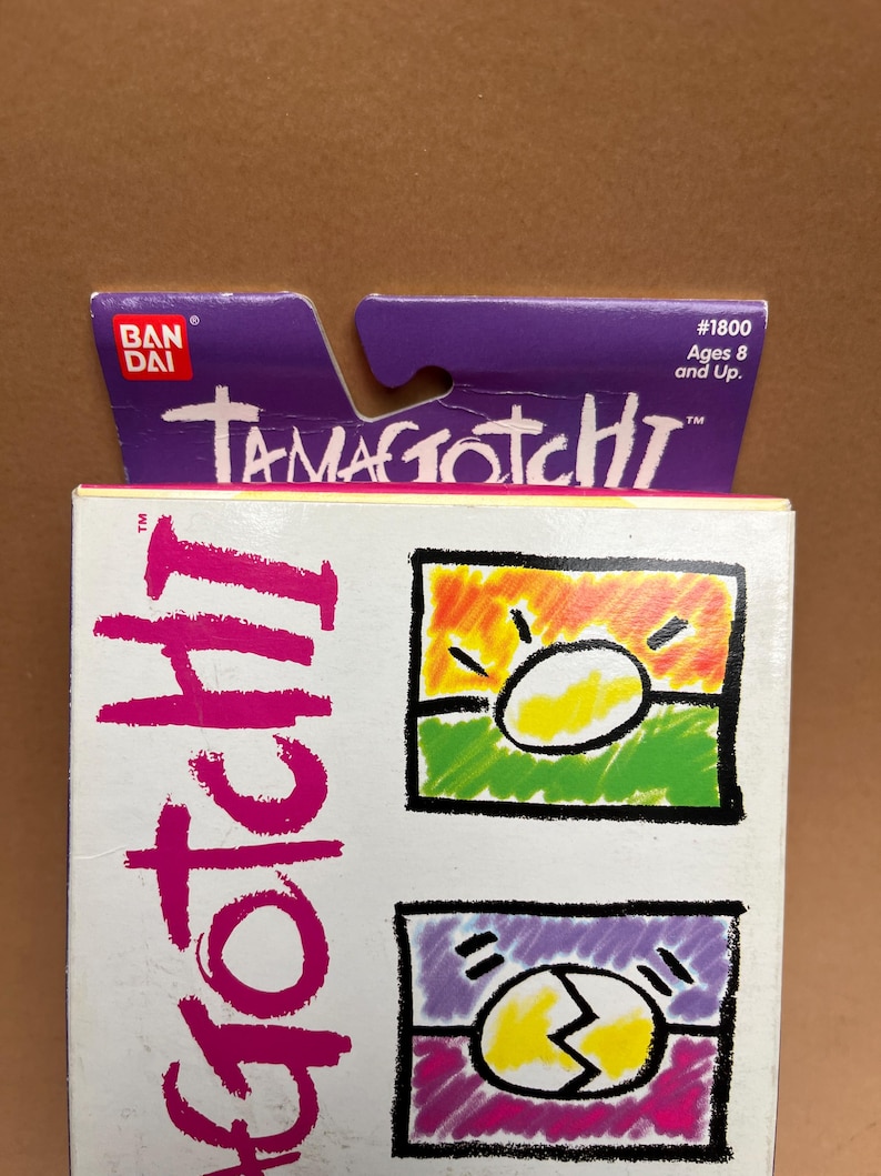 May include: A purple and white box with the text "Tamagotchi" and "#1800 Ages 8 and Up." The box features a cartoon image of a yellow egg with a sun and a cracked egg.