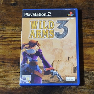 Puede incluir: Caja de videojuego de PlayStation 2 para Wild Arms 3. La caja tiene un borde azul y presenta el título del juego en letras grandes y amarillas. La portada representa a un personaje con ropa morada y azul sosteniendo un arma, sobre un fondo desértico.