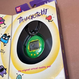 May include: A green Tamagotchi virtual pet on a black chain. The Tamagotchi is in its original packaging with the words "Tamagotchi" and "the original virtual reality pet" printed on the box. The box is yellow with black and red accents. The packaging also includes the text "Bandai 1996-1997. 2 LR44 batteries included."