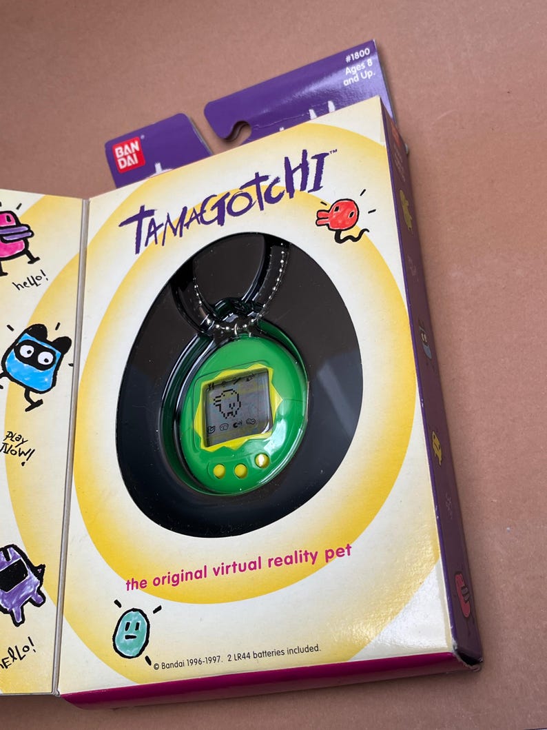 May include: A green Tamagotchi virtual pet on a black chain. The Tamagotchi is in its original packaging with the words "Tamagotchi" and "the original virtual reality pet" printed on the box. The box is yellow with black and red accents. The packaging also includes the text "Bandai 1996-1997. 2 LR44 batteries included."