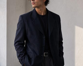 May include: A dark navy blue blazer with a notched lapel and three front buttons, worn over a plain black t-shirt. The blazer features long sleeves with buttoned cuffs, paired with dark grey trousers and a black leather belt with a silver buckle.