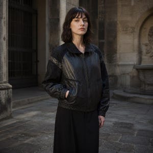May include: A black leather jacket with a zip-up front and a bomber-style design. The jacket has a textured pattern on the shoulders and sleeves. The person is wearing a black skirt. The background is a stone building.