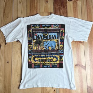 Peut inclure: T-shirt blanc avec un motif graphique coloré représentant un éléphant et le mot "NAMIBIA". Le motif est entouré d'une bordure noire et comprend des motifs géométriques.