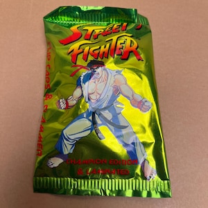 May include: A green and yellow foil pack of Street Fighter trading cards. The pack features a cartoon image of Ryu, a character from the Street Fighter video game series. The text on the pack reads "Street Fighter Champion Edition & Laminated".