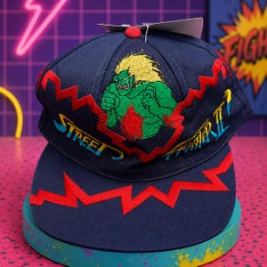 May include: A navy blue baseball cap featuring an embroidered graphic of the character Blanka from Street Fighter II. The cap includes red zigzag stitching across the crown and brim, with the text Street Fighter II embroidered in blue and yellow.