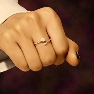 Könnte beinhalten: Ein goldfarbener Ring mit zwei weißen Edelsteinen. Der Ring hat ein zartes, offenes Design, mit einem größeren Edelstein oben und einem kleineren darunter. Der Ring wird an einem Finger getragen und zeigt seine elegante Schlichtheit.