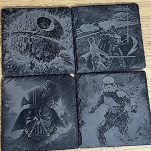 May include: Set of four square slate coasters featuring Star Wars designs. The coasters depict the Death Star, Darth Vader, a Stormtrooper, and a scene with Darth Vader and another character. The images are etched in a grayscale color scheme.