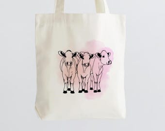 May include: A white tote bag with a pink watercolor background and a black line drawing of three cows.