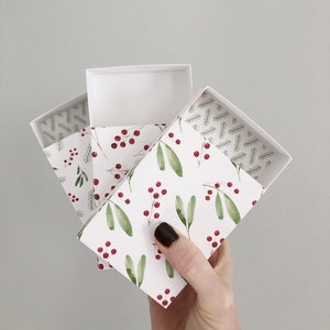 May include: Set of three white gift boxes with a holiday pattern of green leaves and red berries. The boxes are open and stacked on top of each other. The image includes the text "@cuddlebugkids".