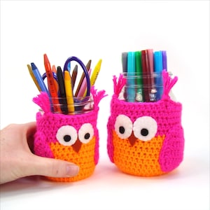 May include: Two crocheted owl pencil holders, one pink and orange and the other pink and orange, with colorful pencils and markers inside.