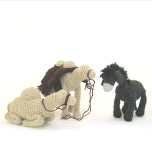 May include: Three crocheted stuffed animals: a camel with a brown saddle, a camel lying down, and a black donkey. The camels are beige and the donkey is black with a grey muzzle.