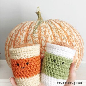 May include: Two crocheted pumpkin spice latte cups with smiling faces. The cups are orange, green, and white. The cups are in front of a large orange pumpkin.